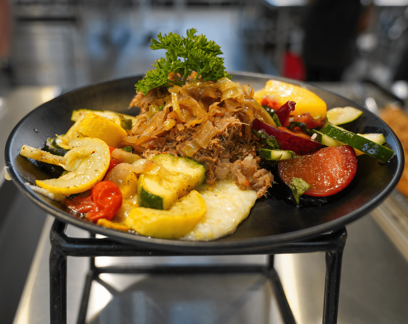 A plate with slow braised apple butter pulled pork, stone ground white corn grits, grilled yellow squash, zucchini, and grape tomatoes.